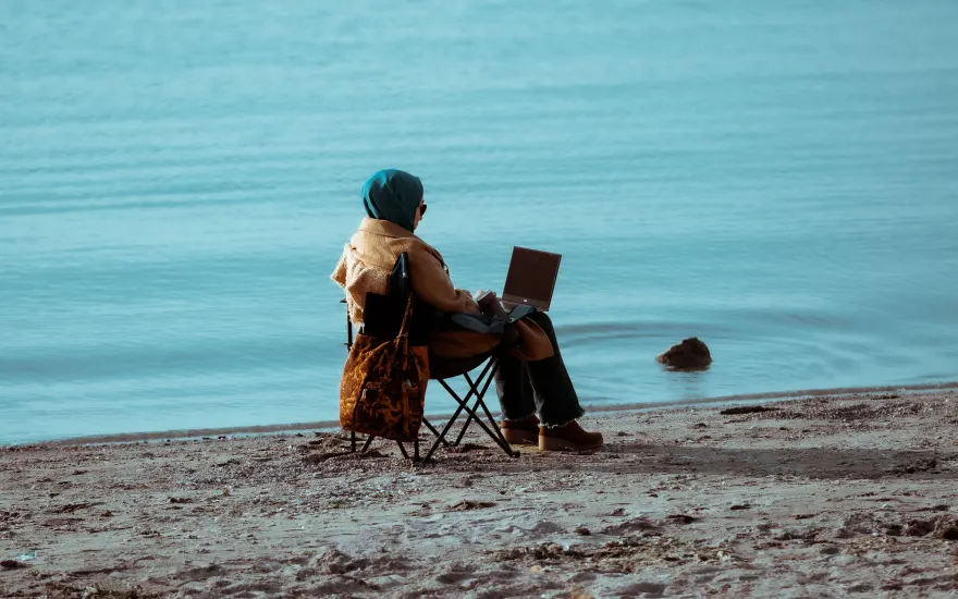 Someone working on a laptop in a folding chair at the edge of the water, a bag propped against the chair and open sand behind them.